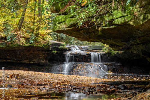 Creation Falls, a waterfall in the Red River Gorge region of Kentucky, cascading over a rocks covered with colorful autumn leaves, is viewed downstream from under an overhanging rock ledge.