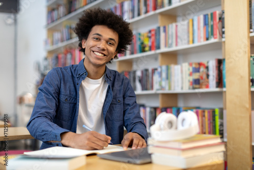 Happy black student guy sits at desk in library, smiling at the camera while writing in copybook, free space