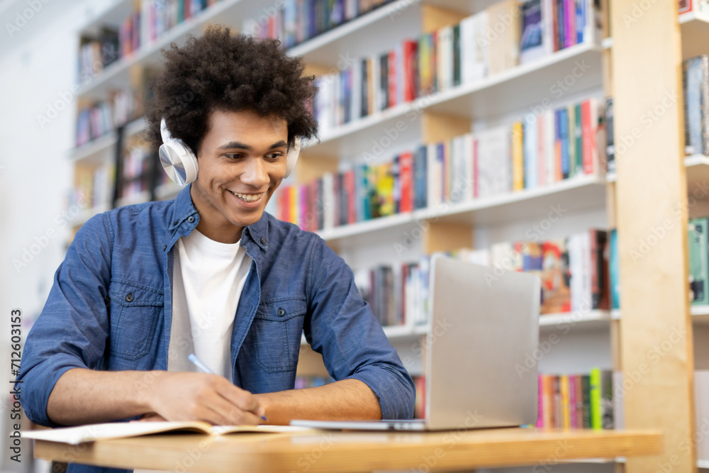 Focused black student guy in headphones taking notes and looking at laptop screen while sitting in university library, scene of concentration and technology-enhanced learning Focused black student guy in headphones taking notes and looking at laptop screen while sitting in university library, scene of concentration and technology-enhanced learning