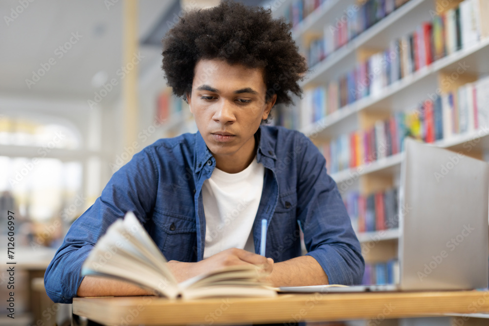 Focused black male student reading book and taking notes, sitting in front of laptop in university library, dynamic combination of traditional reading and technology-enhanced note-taking Focused black male student reading book and taking notes, sitting in front of laptop in university library, dynamic combination of traditional reading and technology-enhanced note-taking