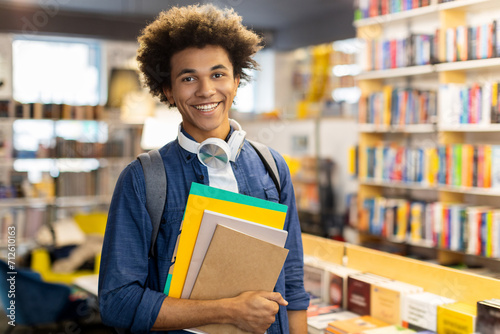 Cheerful african american male student, holds copybooks in library, backpack slung over his shoulders, smiling at camera photo