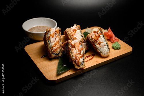 Sushi rolls tempura sandwich on a wooden board with wasabi and ginger, next to a bowl with sauce
