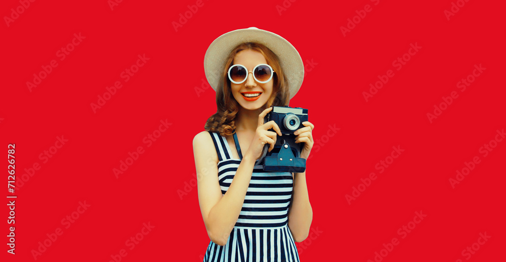 Summer portrait of happy smiling young woman photographer with film camera wearing straw round hat on red studio background