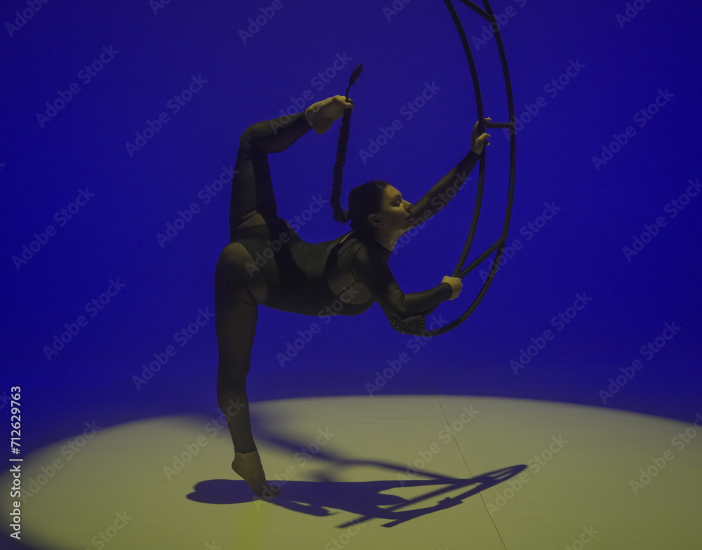 Fototapeta premium Young female aerial gymnast posing near moon on blue yellow studio background. Acrobat in black leotard shows stretching and holds her braid with her foot. Concept of aerial gymnastics, circus show.