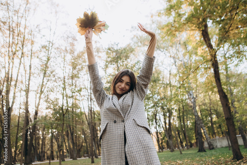 Caucasian woman walking outdoor smiling having fun  portrait of young european lady in warm sunny autumn park season  fall  hold yellow orange red maple leaves  dressed warm coat