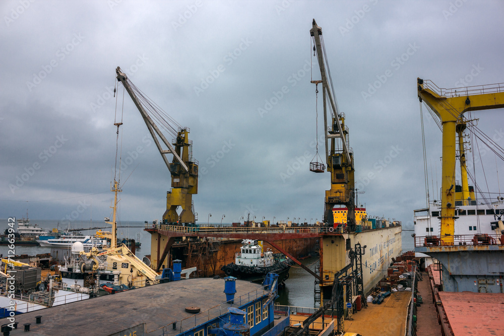 Industrial ship repair yard with floating dock, cranes hoist vessels for maintenance. Maritime engineering, dockyard activity under cloudy sky. Shipping industry, ship refitting, heavy machinery work.