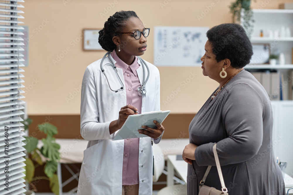 Obraz premium Medium long shot of young African American woman physician in clinic office talking to senior female patient and holding clipboard