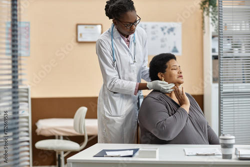 Mature short-haired Black woman patient feeling pain in neck while female doctor conducting medical examination in hospital room photo