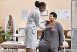 Medium long shot of mature African American woman sitting on examination table in modern clinic while female doctor listening to her heartbeat