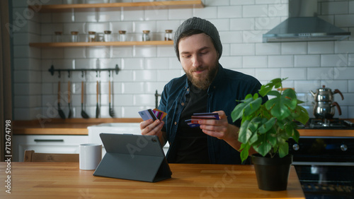 Anxious man with beanie sitting in kitchen at home takes his credit cards out of his pocket and tries them one by one to find the credit card that has not reached its limit photo