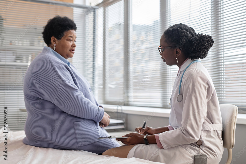 Side view of Black woman doctor sitting in hospital room closely listening to senior female patient complaining of pains Side view of Black woman doctor sitting in hospital room closely listening to senior female patient complaining of pains