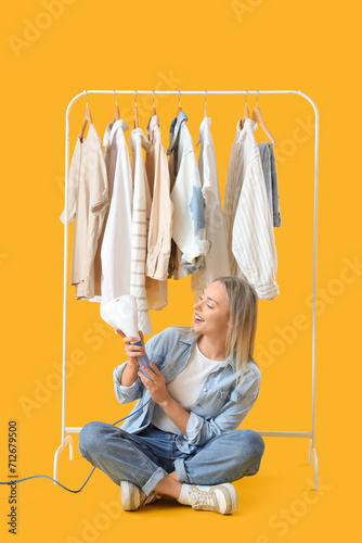 Pretty young woman with modern steamer and clothes on yellow background