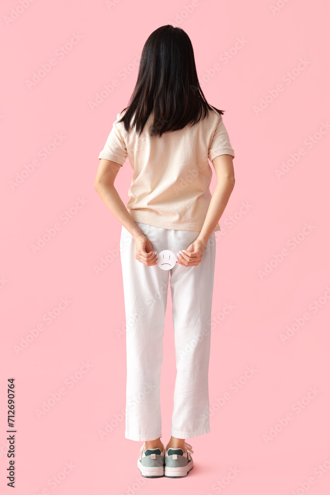 Young woman with hemorrhoids and sad smile on pink background, back view