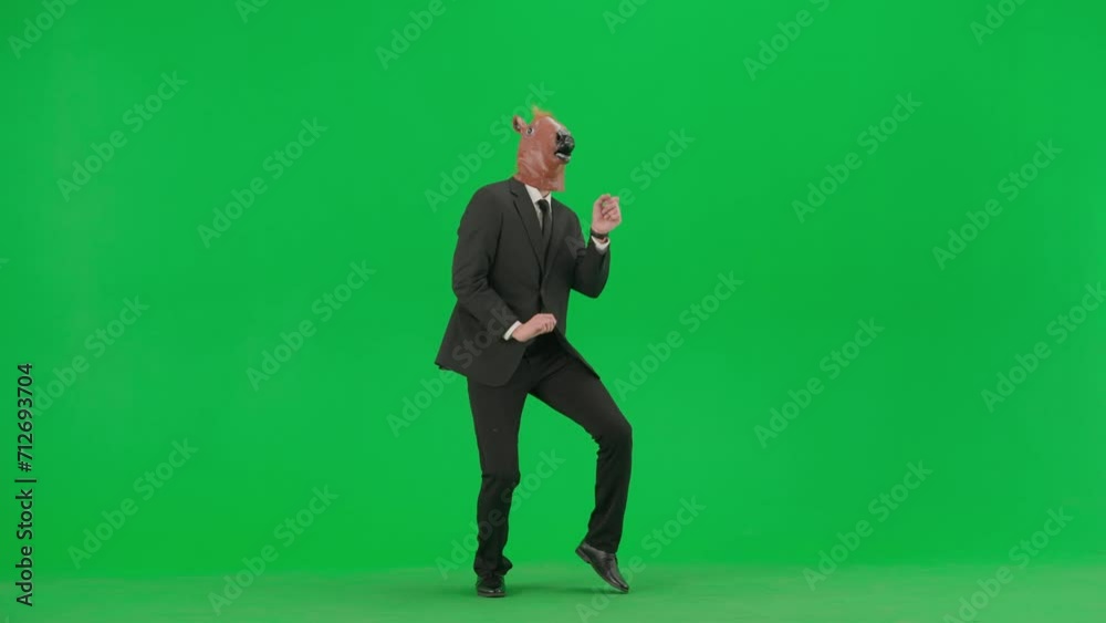A man in a business suit with a horse head mask on a green studio background. Businessman dancing merrily. Hard office work concept. A man in a business suit with a horse head mask on a green studio background. Businessman dancing merrily. Hard office work concept.