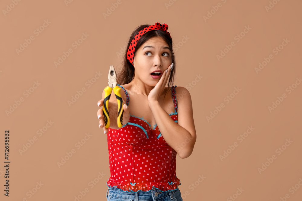 Shocked young Asian pin-up woman with pliers on brown background