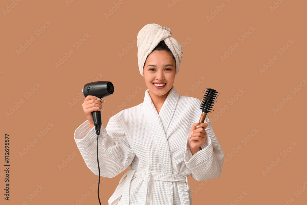 Beautiful young Asian woman in bathrobe with hair dryer and brush on brown background Beautiful young Asian woman in bathrobe with hair dryer and brush on brown background