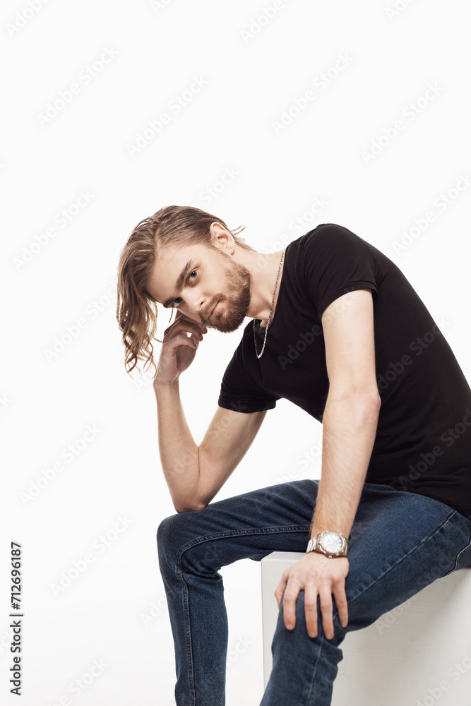 A young guy with a beard sits with his hand resting his cheek on a white background. A young guy with a beard sits with his hand resting his cheek on a white background.