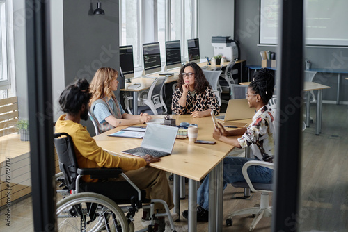 Three multi-ethnic women and indian man in wheelchair having business meeting at desk in office
