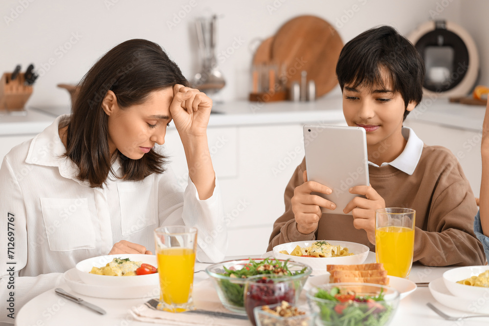 Teenage boy using tablet computer and his upset mother at table during dinner in kitchen. Family problem concept Teenage boy using tablet computer and his upset mother at table during dinner in kitchen. Family problem concept
