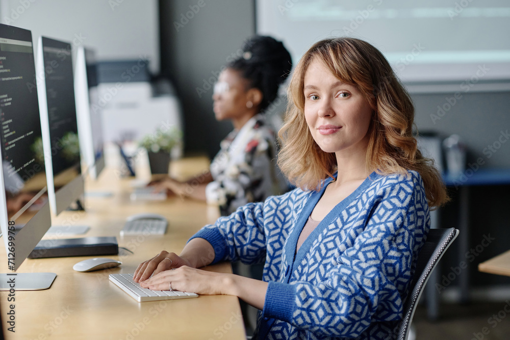 Fototapeta premium Caucasian female coder sitting at her desk working with data on computer and smiling at camera