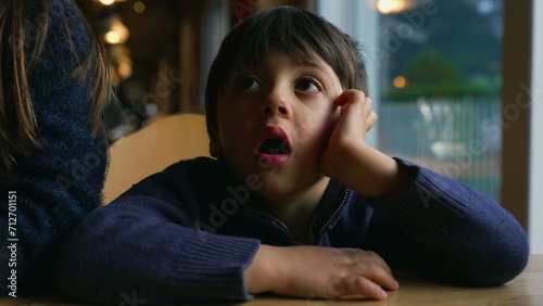 Bored small boy yawning while waiting at restaurant table by window. Child feeling boredom in authentic lifestyle moment awaiting for food to arrive