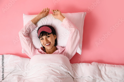 Happy young woman in pajamas with soft pillow and blanket lying on pink background