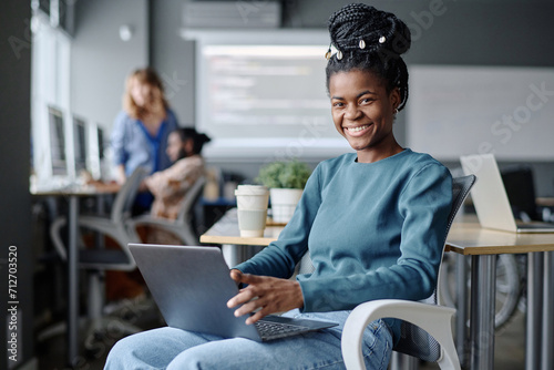 Young female african american coder sitting on chair with laptop on her lap smiling at camera photo