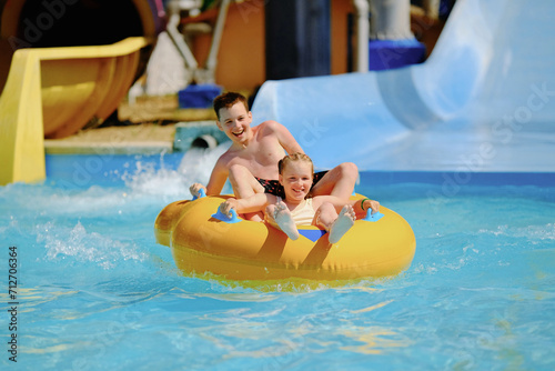 Boy and girl have fun on water slide in outdoor aquapark. Little children floating on yellow inflatable raft.