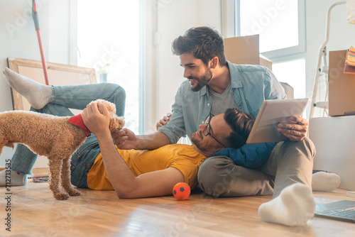 Happy homosexual couple sitting on floor and enjoying new home with poodle dog.
