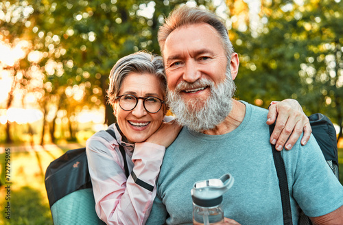 Portrait of active sincere carefree smiling happy modern gray-haired couple shouldering bag with sports mats, man holding water. Sports jogging, training in the park in the morning. © HBS