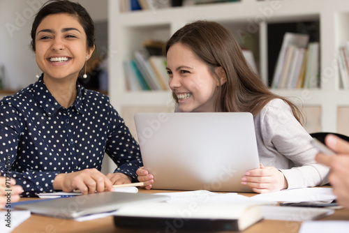 Excited multiethnic girls sit at desk in classroom laugh brainstorming learning preparing for exam, happy smiling multiracial female students have fun gather work on shared project or study together