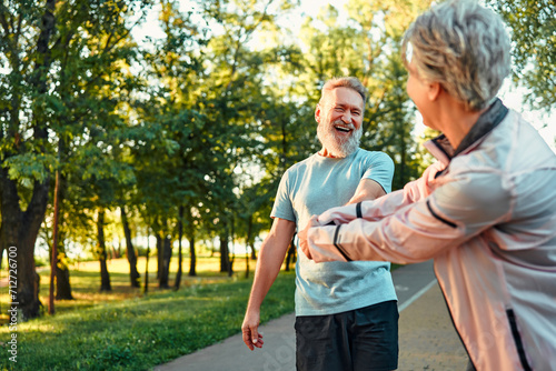 Happy couple outdoors in the park before a morning jog. An adult woman jokingly pulls her friend to run, and he stops and laughs.Seniors lifestyle. Copy space. Outdoors activities. photo