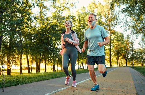 Fototapeta Naklejka Na Ścianę i Meble -  Happy and healthy lifestyles. Caucasian aged people with sincere smile on faces enjoying outdoors cardio at green park. Active male and female pensioners in sport outfits spending time for workout.