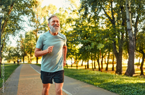 Physical activity on retirement. Happy aged man in sport clothes running alone at green summer park during morning time. Smiling caucasian pensioner having regular workout for fit and healthy body.