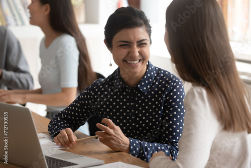 Overjoyed multiracial millennial female students sit at desk in classroom laugh working on educational project together, smiling multiethnic girls have fun joke brainstorming cooperating at lesson
