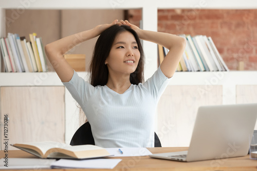 Dreamy millennial ethnic girl sit at desk studying look in distance thinking or visualizing, pensive Asian female student lost in thoughts distracted from learning dreaming relaxing at workplace photo