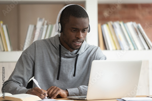 Concentrated biracial male student in Bluetooth headphones look at laptop make notes taking online training distant course, focused african American guy in earphones study watching webinar on computer photo