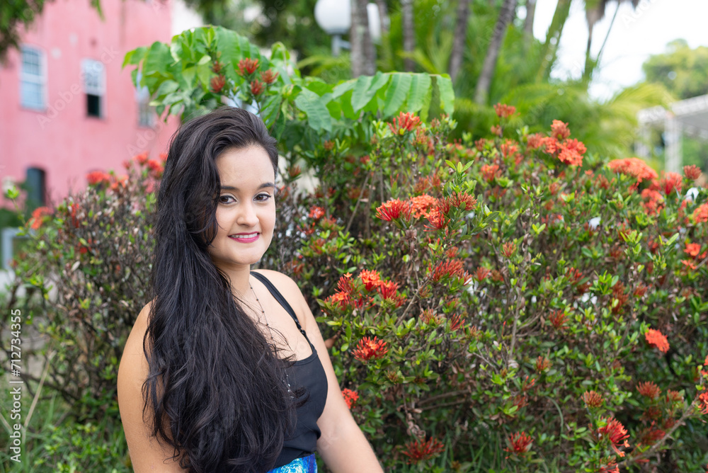 Portrait of a beautiful young woman, with long black hair, looking at the camera with flowers in the background. Portrait of a beautiful young woman, with long black hair, looking at the camera with flowers in the background.