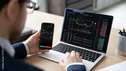 Tableau sur toile Male trader sits at desk hold smart phone with stock market data, using laptop display graph and chart, analyzing investment, check trading stocks information, view over shoulder