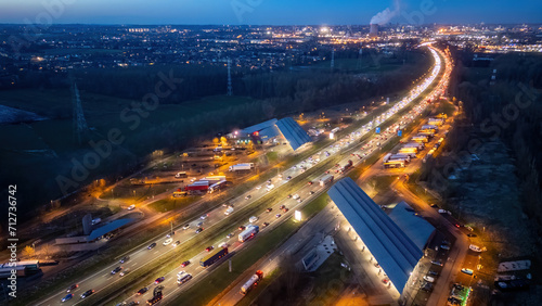 This aerial image captures the bustling E19 highway as it courses near Halle, Brussels, during evening rush hour. The scene is alive with the glow of headlights and tail lights, illuminating the