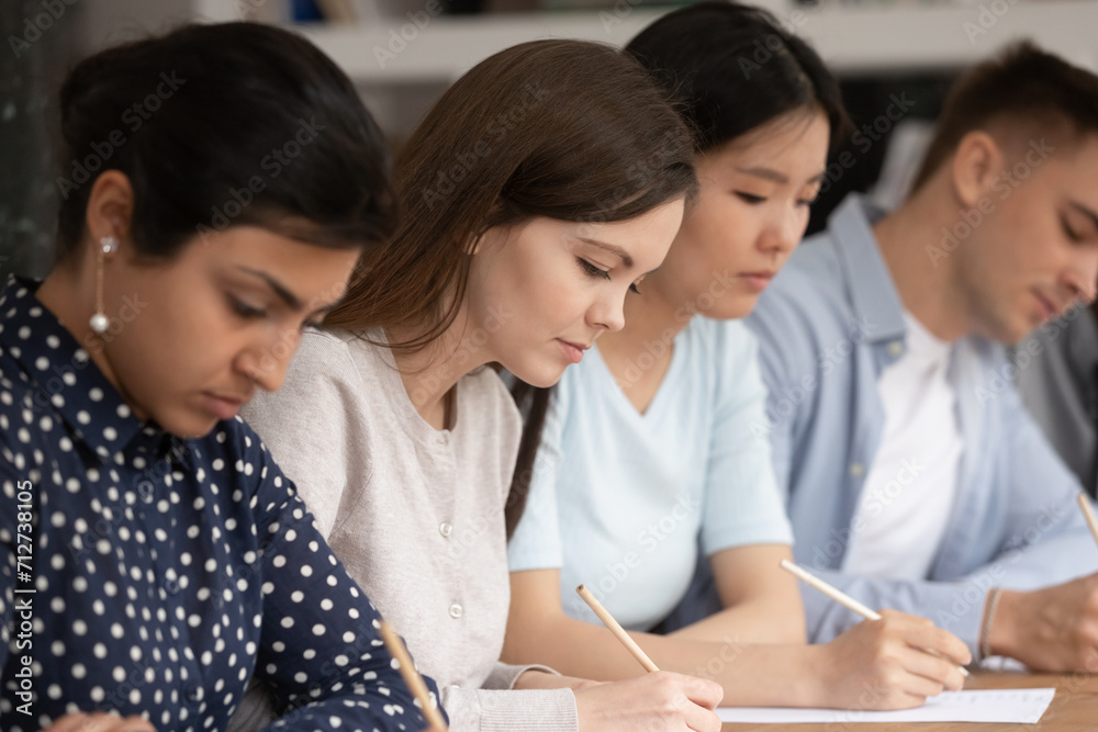 Concentrated multiracial young people sit in row at table busy writing