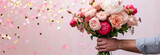 cropped image of a man’s arm holds out a beautiful bouquet of roses, peonies and ranunculuses isolated on light pink pastel background with round golden and pink blurred confetti 