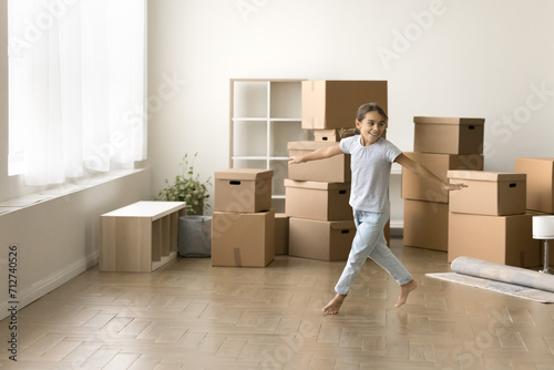 Happy active kid girl running past stacked moving boxes in empty room, dancing, hopping, celebrating relocation into new apartment, flat, house, having fun, enjoying being at new home