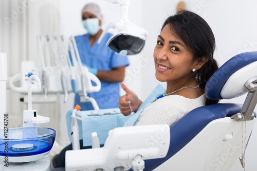 Happy woman sitting in dental chair after teeth cure giving thumb up