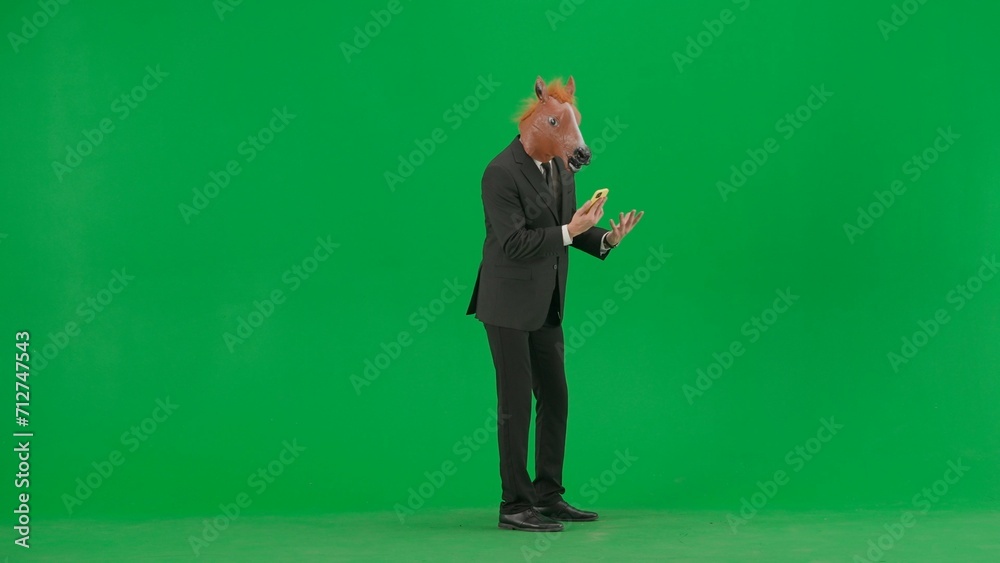 A man in a business suit with a horse head mask on a green studio background. A businessman is talking on a smartphone, gets angry and shouts into the phone. Hard office work concept. A man in a business suit with a horse head mask on a green studio background. A businessman is talking on a smartphone, gets angry and shouts into the phone. Hard office work concept.