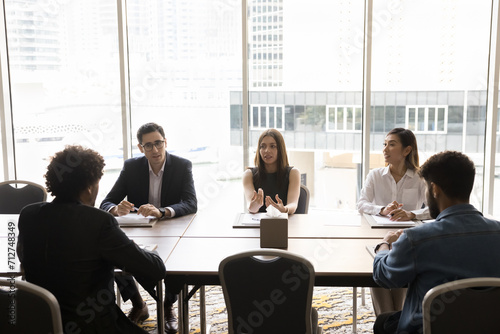 Young business leader woman talking to diverse team of colleagues, speaking at large meeting table, explaining work tasks, motivating workers. Corporate coach giving seminar to interns