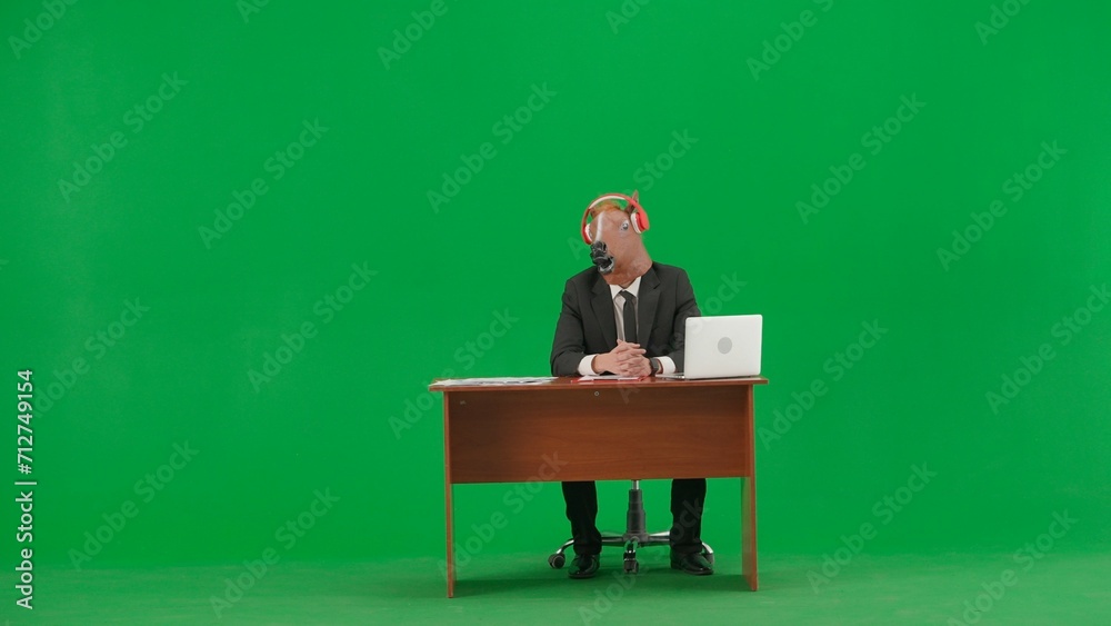 Man in business suit with horse head mask on studio green background. Businessman sitting at desk, listening to music with big red headphones. Concept of heavy office work. Man in business suit with horse head mask on studio green background. Businessman sitting at desk, listening to music with big red headphones. Concept of heavy office work.