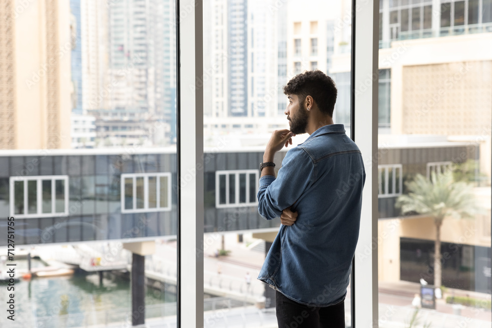 Thoughtful serious handsome young Arab man in casual informal clothes standing at large window, looking at city buildings view, touching chin, thinking on apartment buying, real estate development