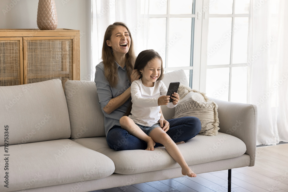 Little pretty daughter sits on laughing loving mothers laps, holds smartphone, having fun, make photos, play new game, enjoy mobile application, talk to friend using videocall app on modern cellphone