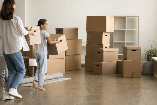 Pretty daughter kid and young mom carrying moving boxes in new apartment, walking to heap of stacked cardboard containers, enjoying relocation activity after buying house